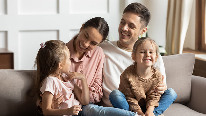 Giovane papà spinge per gioco un carrello bagagli con sopra mamma, figlio e figlia. Tutti indossano abiti molto sportivi e colorati, la famiglia è pronta per iniziare la vacanza 